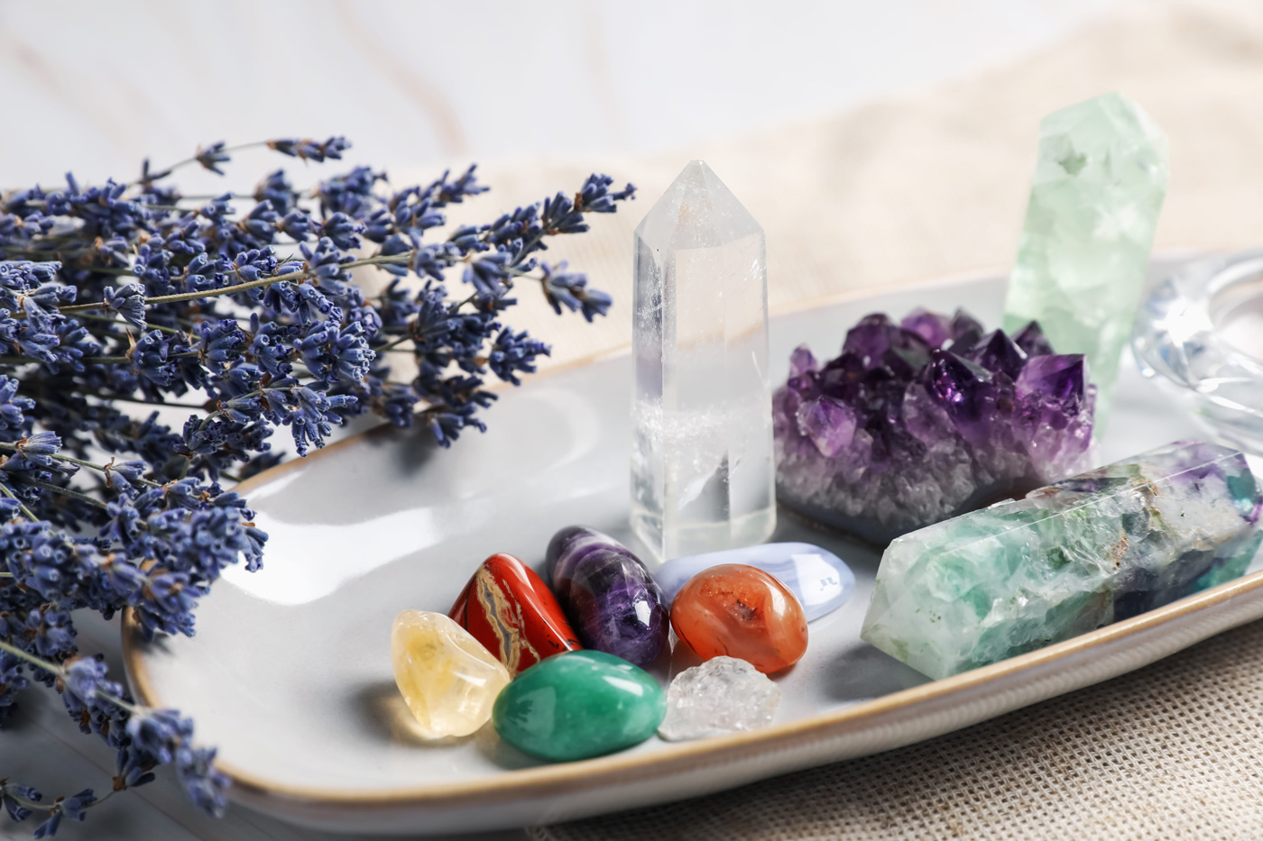 A clear quartz crystal, an amethyst geode, several vibrant gemstones, and dried lavender on a table together.